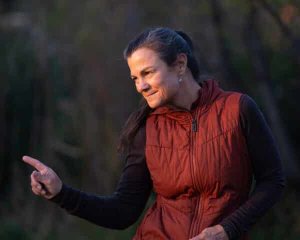 A woman with long brown hair pulled back, wearing a maroon vest over a dark shirt, smiles and points with her right index finger in an outdoor setting, perfect for showcasing hunting property or land for sale with a blurred forest background.