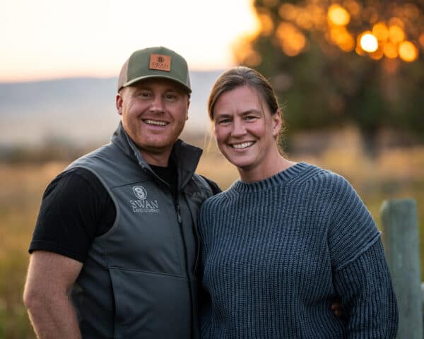 A smiling man in a vest and cap stands beside a smiling woman in a sweater outdoors at sunset, with blurred trees and fields on a beautiful cattle ranch in the background.