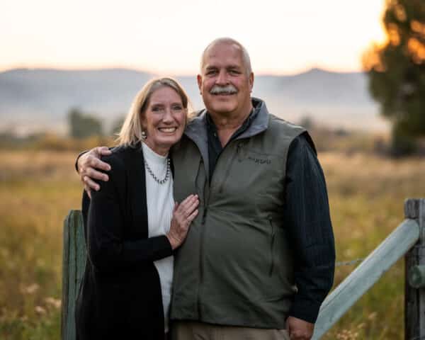 An older couple stands smiling together outdoors by a wooden fence on their cattle ranch, arms around each other. The background features a field, trees, and hills at sunset.