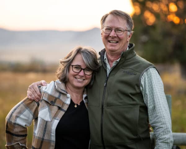 A smiling couple stands outdoors, with the man’s arm around the woman’s shoulders. They wear casual jackets and glasses, set against a blurred natural landscape at sunset—perfect for showcasing a hunting property or cattle ranch.