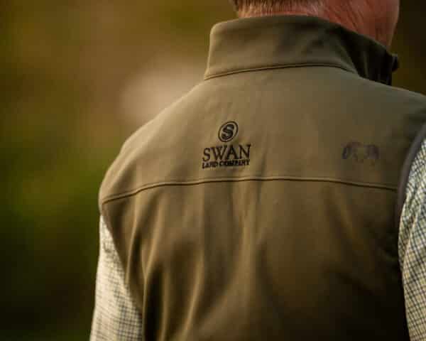 A close-up of the back of a person wearing a green vest with the Swan Land Company logo, highlighting expertise in ranch for sale and recreational land. The person stands outdoors, showing only their upper back and part of their shirt.