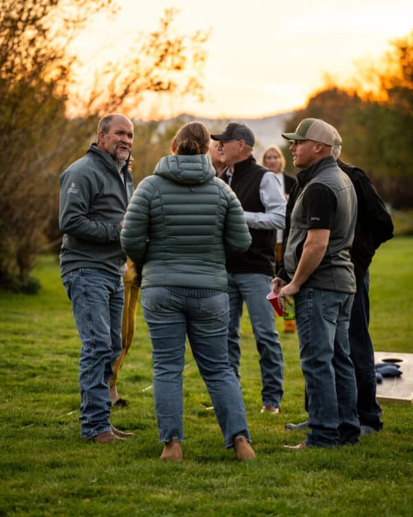 A group of people in casual outdoor clothing stand on grass, talking together at sunset. Trees and soft sunlight are in the background, creating a warm, relaxed atmosphere perfect for exploring recreational land or a cattle ranch.