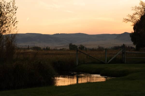 A quiet stream reflects the orange glow of sunset, flowing through grassy fields with a wooden gate and distant rolling hills—an idyllic setting for a cattle ranch or land for sale.