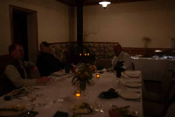 Three men sit around a dimly lit dining table with plates of food, drinks, and candles. The room's stone and wood decor evokes the rustic charm of a cattle ranch, with flower centerpieces and serving dishes on a side table in the background.