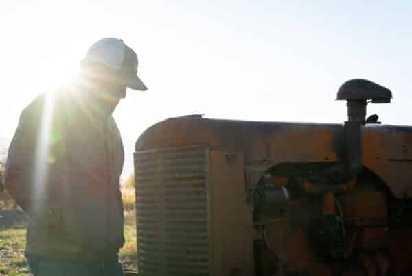A person in a baseball cap and jacket stands beside an old, rusty tractor at sunrise or sunset, with sunlight creating a bright lens flare—suggesting the promise of ranch for sale on open land.