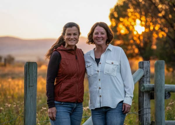 Two women stand side by side, smiling, in front of a wooden fence on a hunting property at sunset. One wears a red vest and black sleeves, the other a white shirt. The background features trees and warm sunlight.