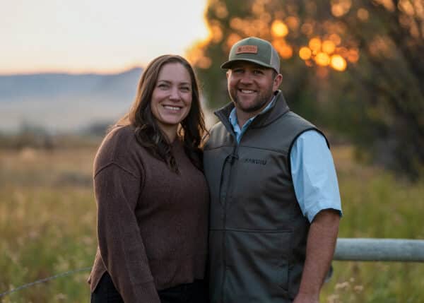 A smiling couple stands outdoors by a fence at sunset on recreational land, with trees and fields in the background. The woman wears a brown sweater, and the man wears a vest, blue shirt, and tan cap.