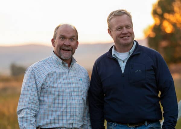 Two men stand outdoors in a field at sunset, possibly surveying a hunting property. One wears a plaid shirt with a surprised expression; the other, in a navy jacket, smiles. Trees and hills appear in the blurred background.