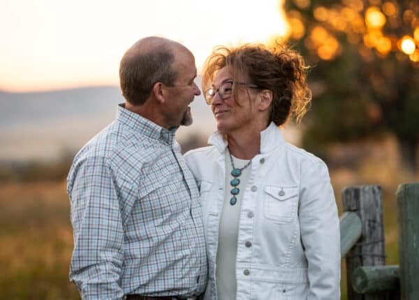 A middle-aged couple stands close together outdoors at sunset, smiling warmly at each other on scenic recreational land. The man wears a plaid shirt and the woman wears glasses, a white jacket, and a turquoise necklace. Trees and soft sunlight frame the scene.