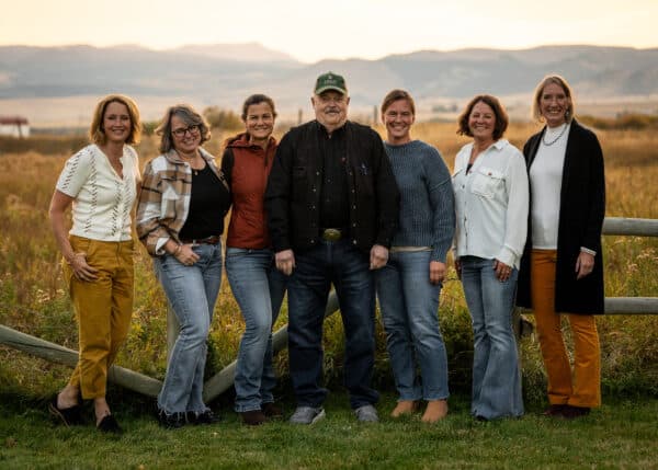 Seven adults stand side by side, smiling, in front of a wooden fence on a cattle ranch with grassy fields and mountains in the background at sunset. They are casually dressed and appear relaxed and happy.