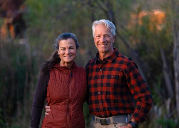 A smiling middle-aged couple stands outdoors on a scenic land for sale, with the man in a red and black plaid shirt and the woman in a maroon vest. They are close together, surrounded by greenery in soft evening light.