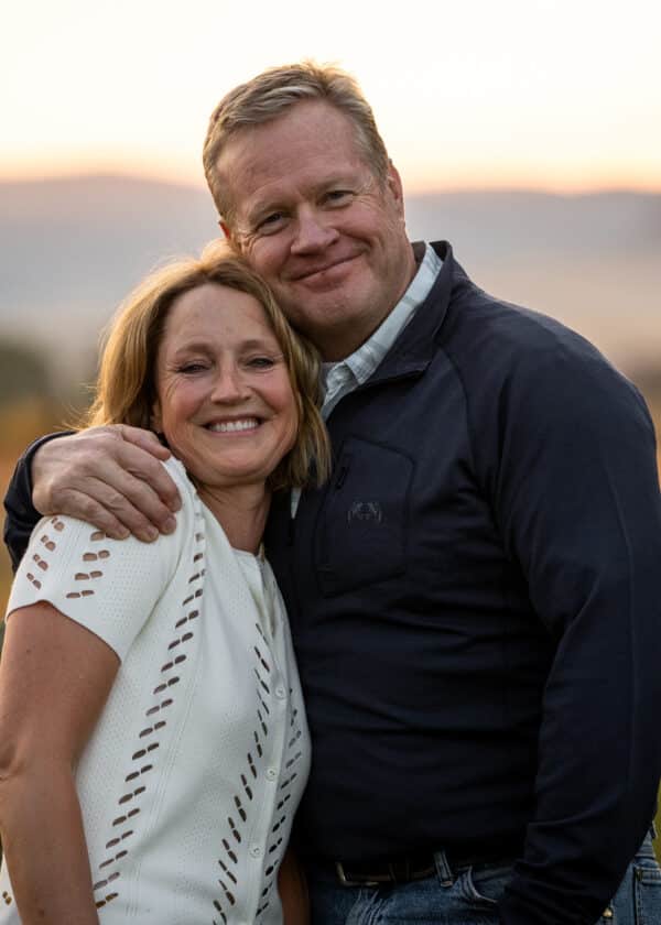 A smiling middle-aged couple stands close together outdoors at sunset on beautiful recreational land, with the man’s arm around the woman’s shoulders. Both appear happy and relaxed, with soft natural light in the background.