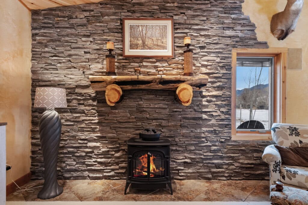 A cozy living room on a cattle ranch features a stone accent wall, wood stove with a fire, rustic wooden mantle, candles, framed artwork, modern floor lamp, snowy window view, and part of a patterned armchair.