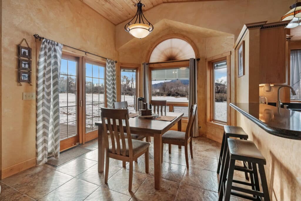 Cozy dining area with wooden table and chairs next to large windows and glass doors, offering scenic views of a snowy landscape and distant mountains—perfect for a hunting property or recreational land retreat, with tiled floor and warm, natural light throughout.
