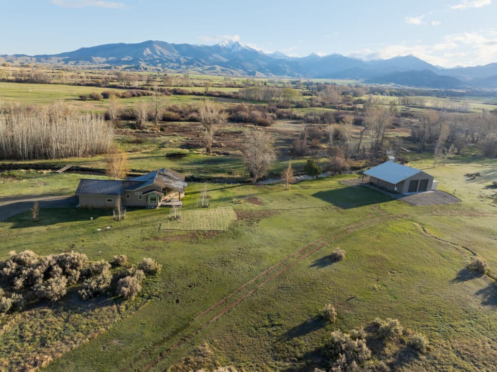 Aerial view of a rural landscape featuring a house, large barn, grassy fields, scattered trees, and distant mountains under a partly cloudy sky—ideal ranch for sale or hunting property.