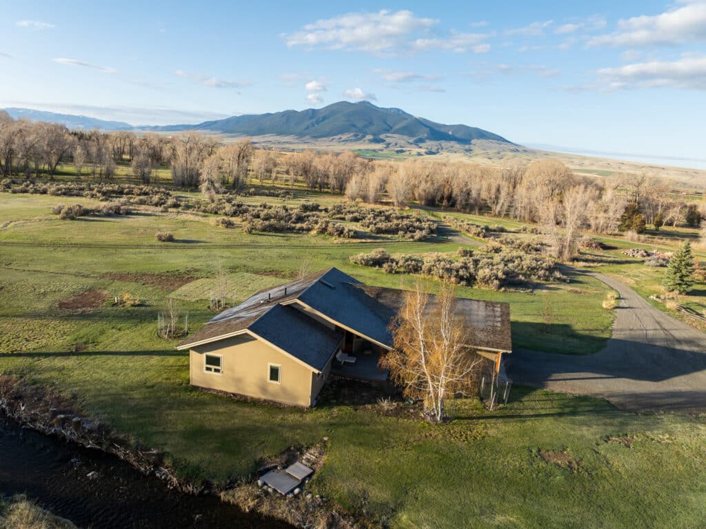 A small tan house with a dark roof sits beside a creek in a grassy field on recreational land, surrounded by trees, with a mountain range in the background under a blue sky with some clouds.
