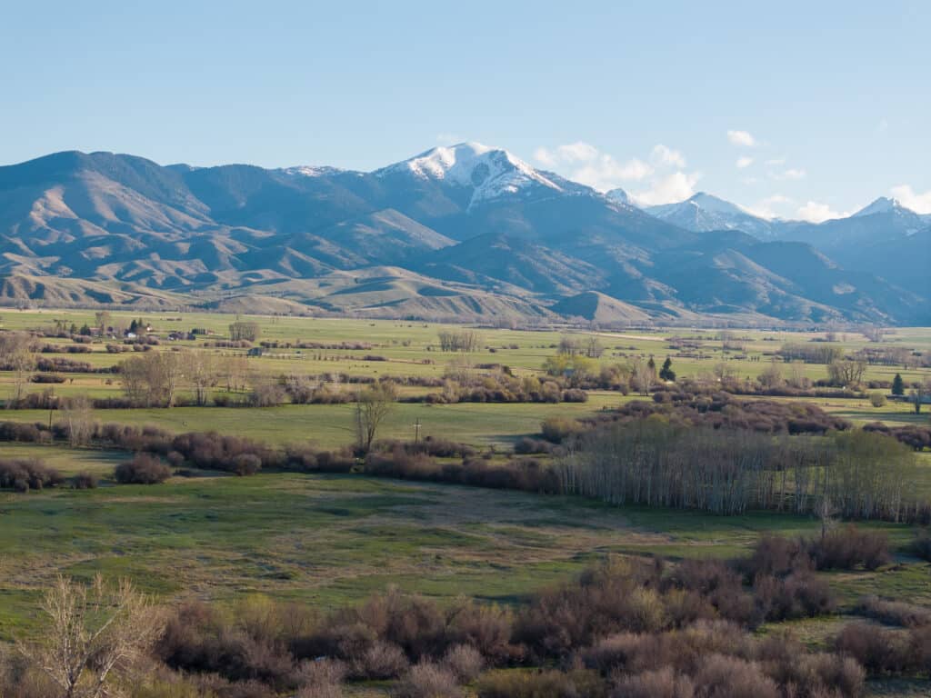 Wide view of a grassy, green valley with scattered shrubs and trees, set against rolling hills and snow-capped mountains under a clear blue sky—ideal recreational land or hunting property.