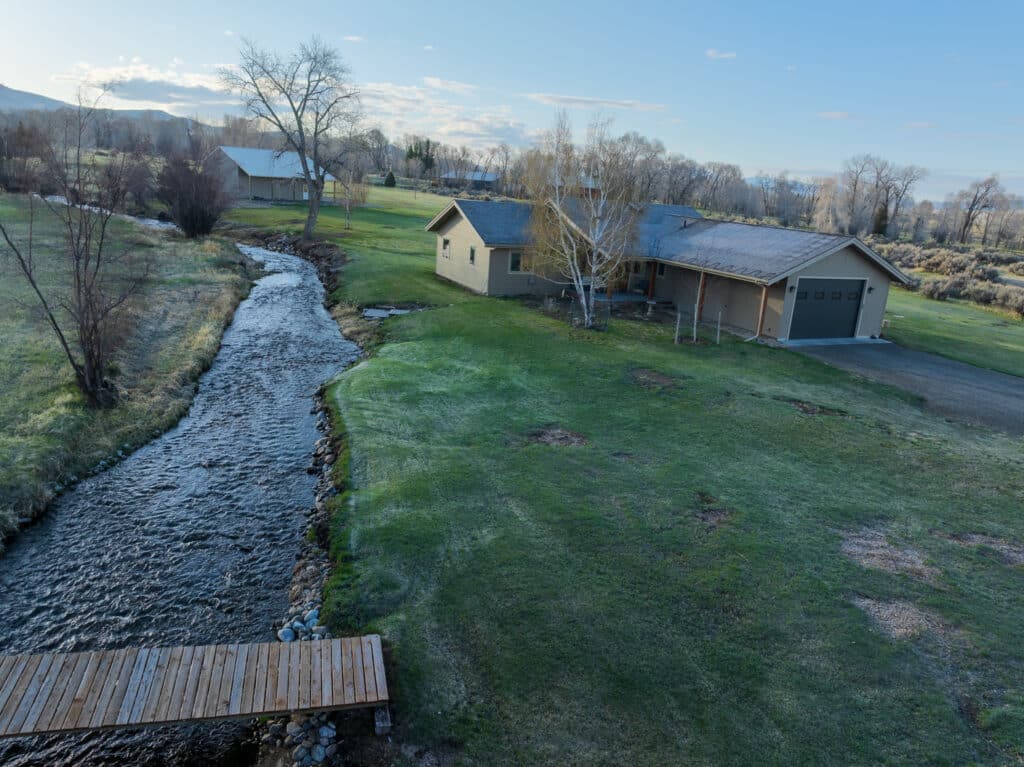 A small creek flows beside a house with a wooden footbridge crossing the water, ideal for recreational land. The grassy yard is surrounded by bare trees, and another building is visible in the background under a clear blue sky.