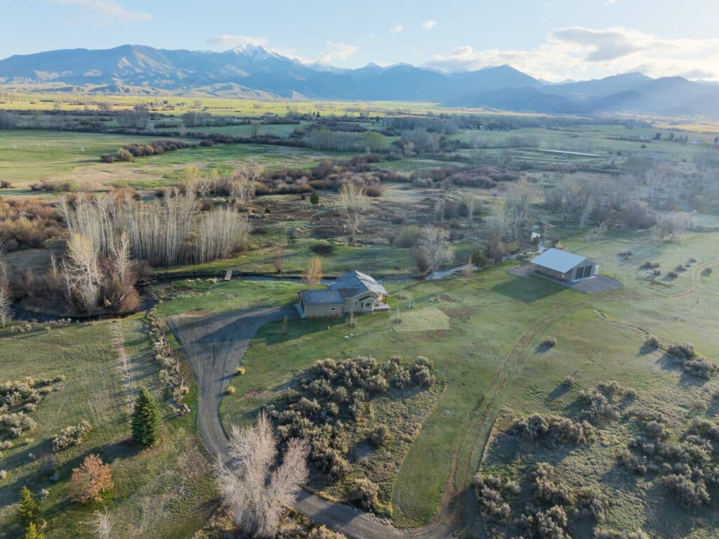 Aerial view of a ranch for sale featuring a house and large shed on a rural property, surrounded by fields, trees, and mountains under a blue sky.