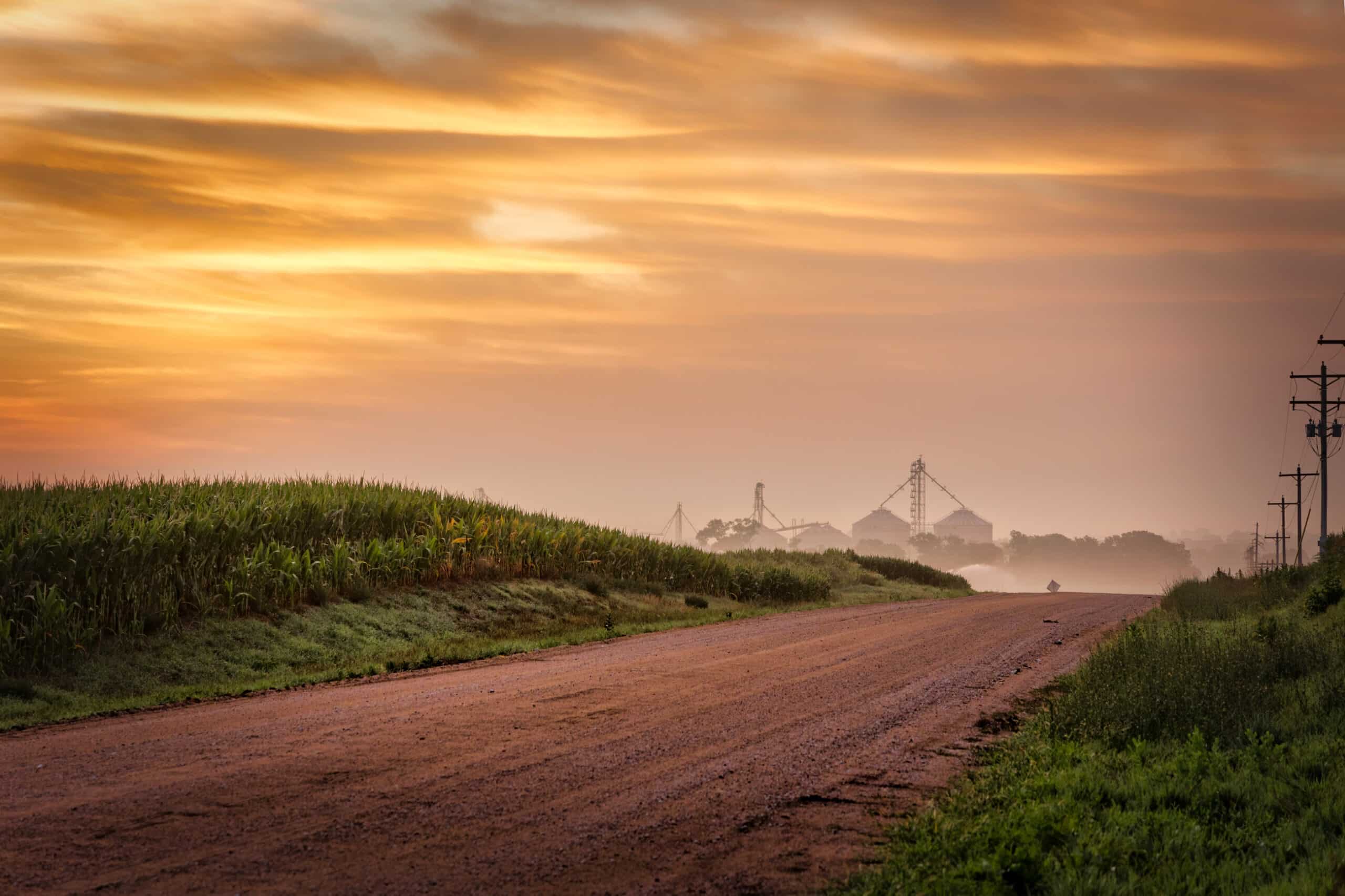 A rural dirt road stretches past green fields under a golden-orange sunrise, with distant silos and utility poles visible through morning mist—perfect scenery for a recreational land or hunting property.