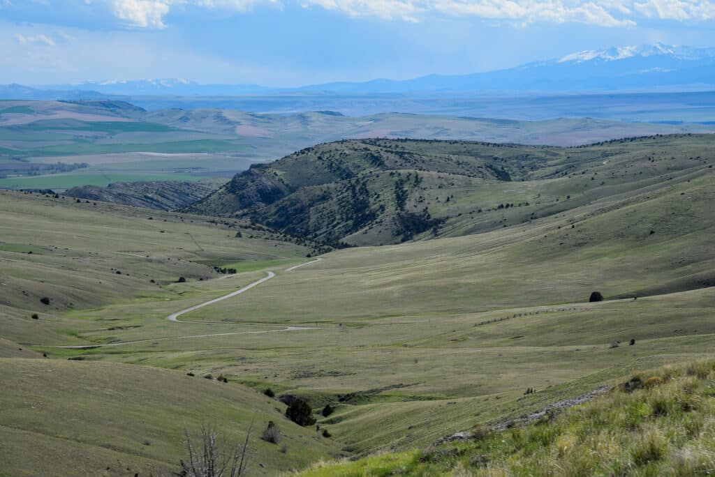 A winding road runs through rolling green hills under a partly cloudy sky, with distant snow-capped mountains on the horizon—perfect recreational land or ideal for a hunting property.