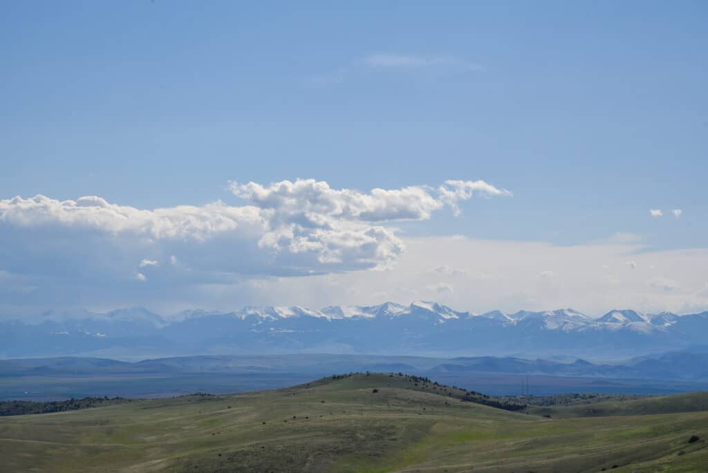 A wide view of rolling green hills, ideal as recreational land, with snow-capped mountains and a blue sky with scattered clouds in the background.