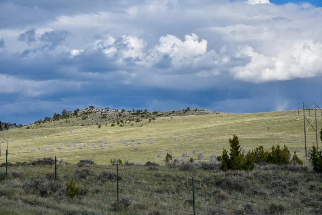 A grassy hillside with scattered shrubs and small trees under a cloudy sky. A wire fence runs along the foreground, and a power line is visible on the right side—ideal as recreational land or hunting property.