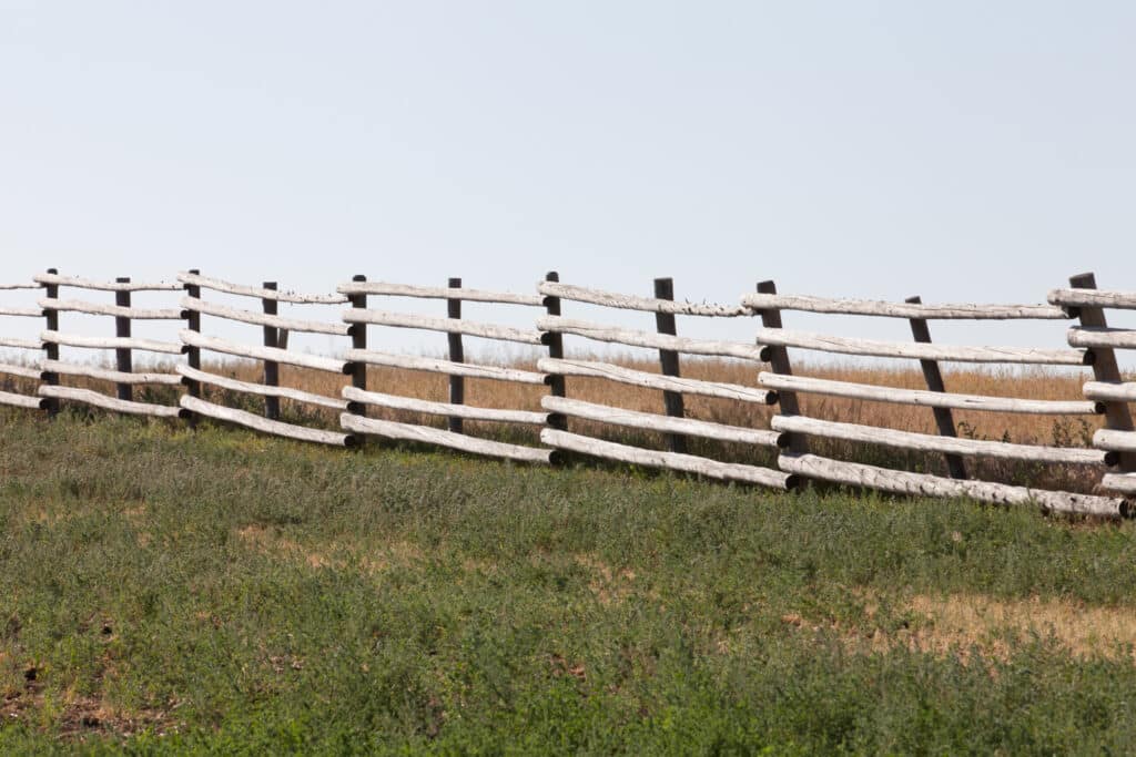 A wooden split-rail fence runs across a grassy field on this scenic hunting property, with some rails leaning unevenly. The sky above is clear and pale blue.