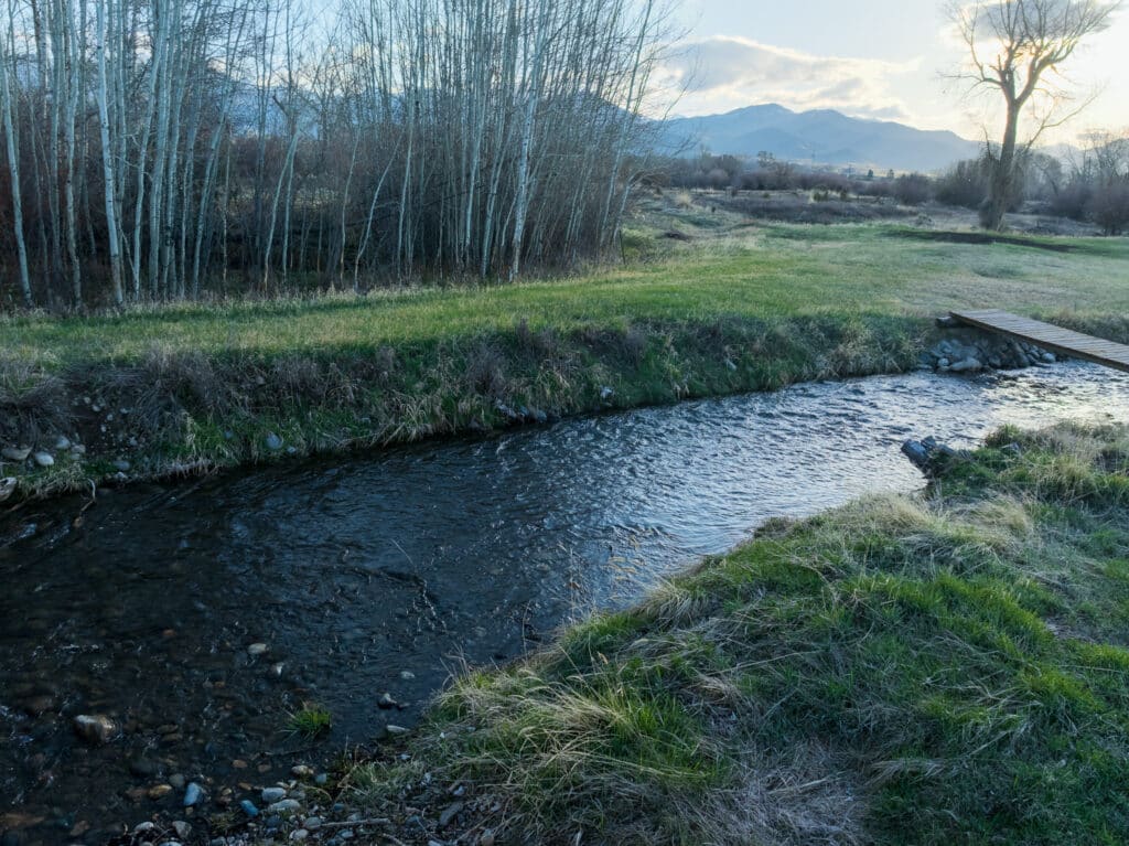A small creek winds through grassy recreational land with leafless trees on one side and a wooden footbridge. Mountains and a cloudy sky are visible in the background at sunset, highlighting this potential hunting property.