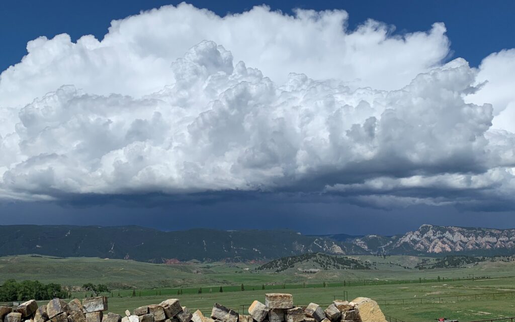 Large, fluffy white clouds dominate the sky above green fields and distant mountains, hinting at prime recreational land with a line of rocks in the foreground under bright daylight.
