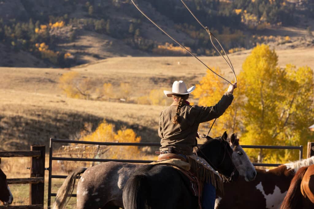 A person wearing a cowboy hat rides a horse and swings a lasso in a fenced area on a cattle ranch, with autumn trees and rolling hills in the background.