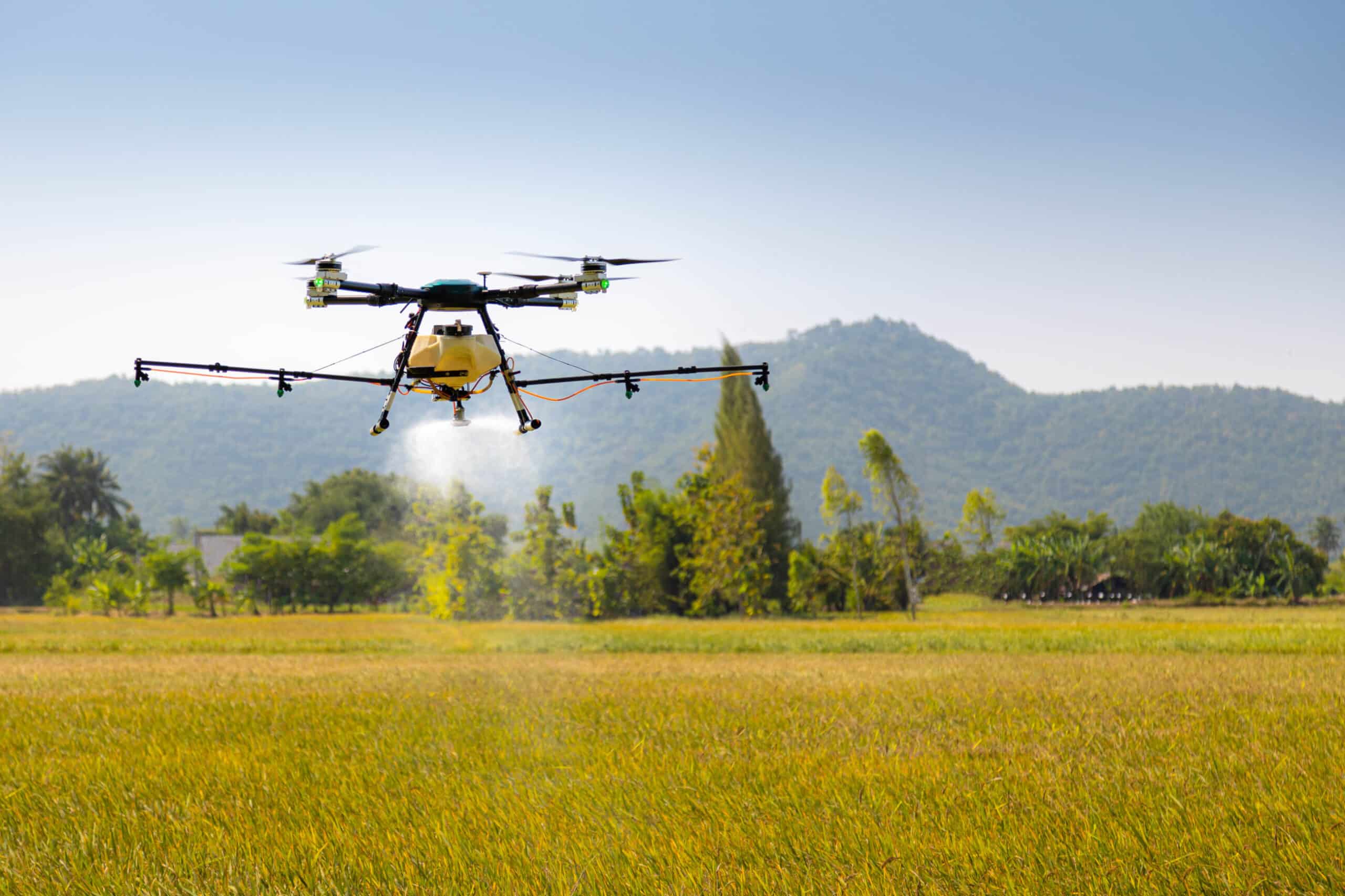 A drone flies low over a field on recreational land, spraying crops, with green hills and trees in the background under a clear blue sky.