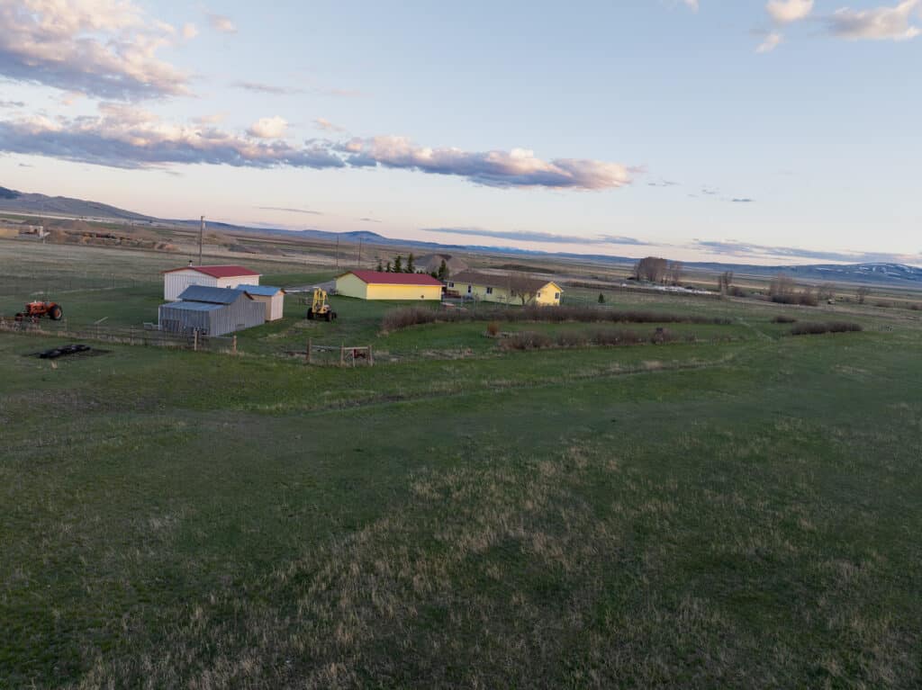 A rural landscape with a few farm buildings, some machinery, and open grassy fields under a partly cloudy sky at sunrise or sunset—ideal as a hunting property or cattle ranch. Mountains rise in the distance. Land for sale.