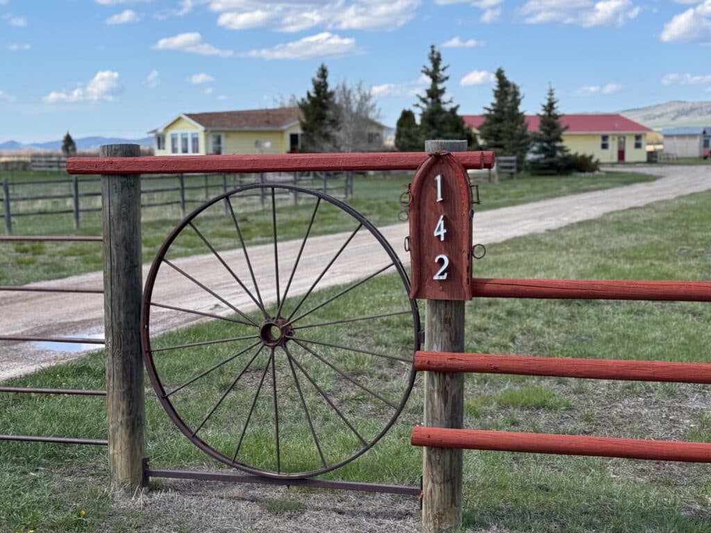 A rustic ranch gate with a large wagon wheel design and a wooden sign displaying the number 142 welcomes you to recreational land. A gravel driveway leads to yellow houses and trees under a blue sky with clouds.