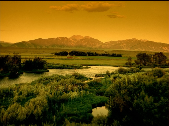 A river winds through lush green vegetation and mountain ranges under an orange-tinted sky, offering stunning views—ideal recreational land for sale.