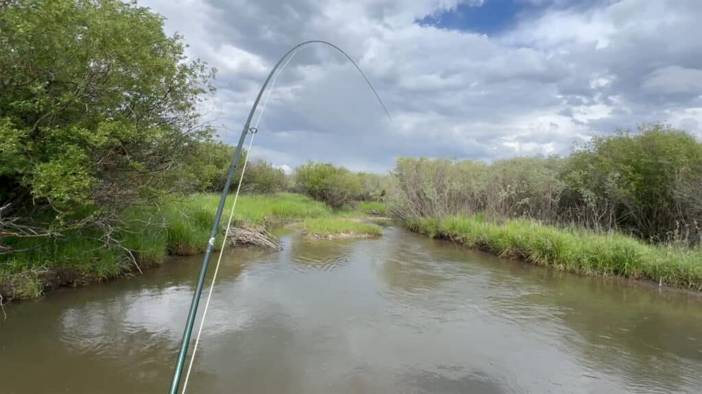 A fishing rod bends over a calm, narrow stream surrounded by green grass and bushes under a partly cloudy sky—picture-perfect scenery on a ranch for sale.