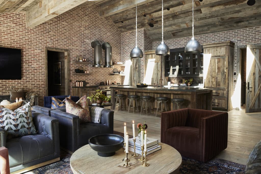 A rustic kitchen and living area with exposed brick walls, wooden ceiling, metal pendant lights, dark sofas, patterned pillows, an armchair, a round coffee table with candles, and a large island with barstools—perfect for your ranch for sale.