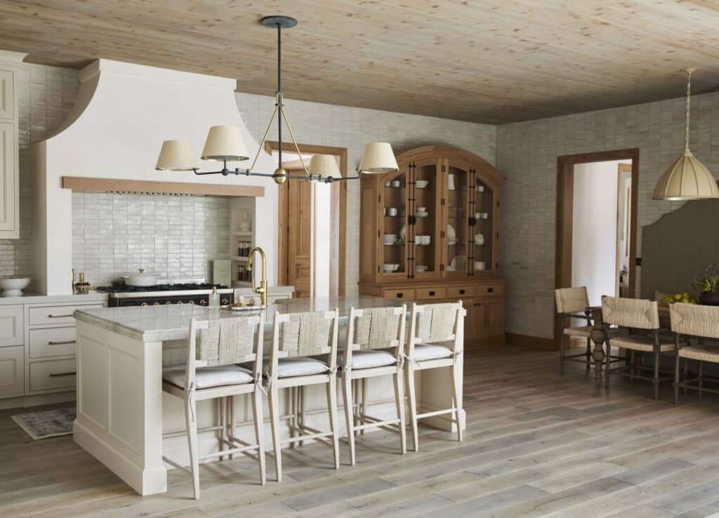 A spacious kitchen with a large island and four white barstools, light wood floors, white cabinetry, tiled backsplash, a wood hutch, and a dining table—perfect for gatherings after a day exploring hunting property or land for sale.