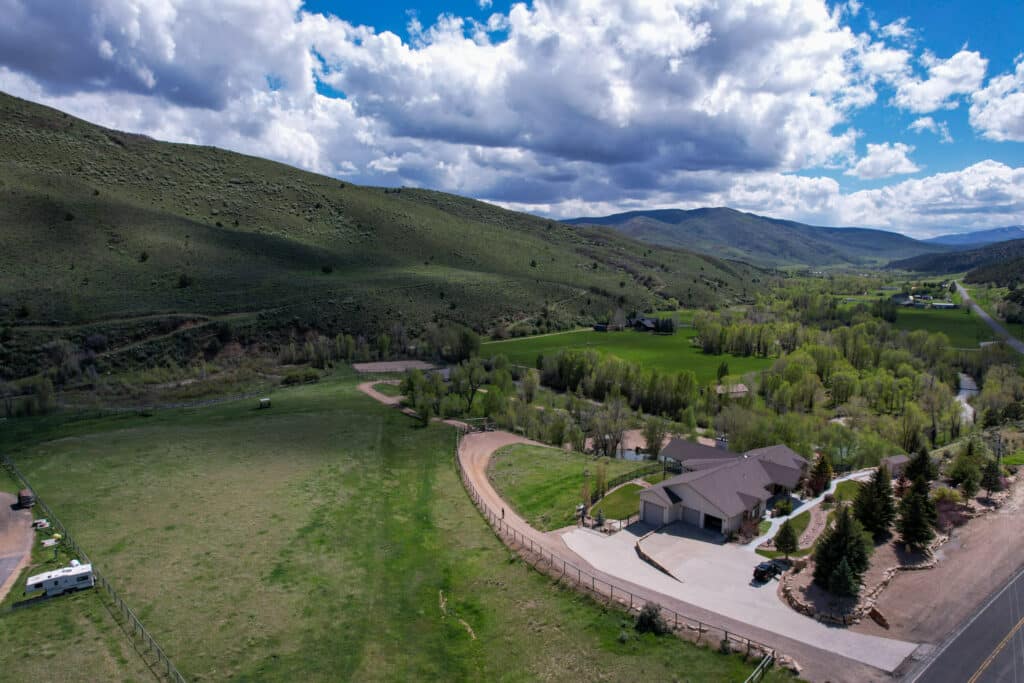 Aerial view of a rural landscape featuring green hills, scattered trees, a house with a driveway, fenced fields, and a country road—ideal as a cattle ranch or hunting property under a partly cloudy sky.