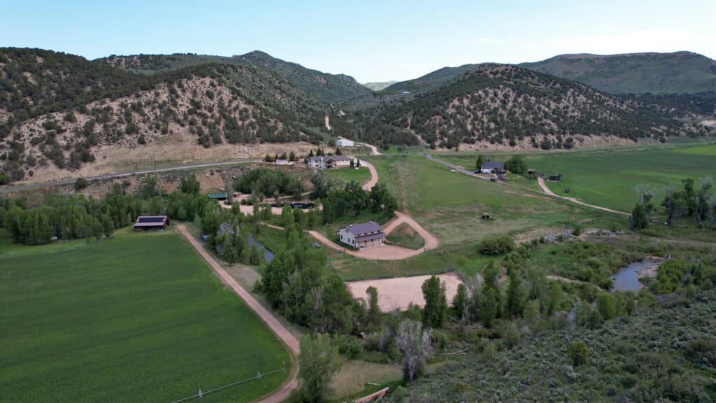 Aerial view of a rural landscape with scattered buildings, green fields, dirt roads, and tree-lined hills under a clear blue sky. A small creek runs through the lower right area—ideal recreational land or cattle ranch with land for sale.