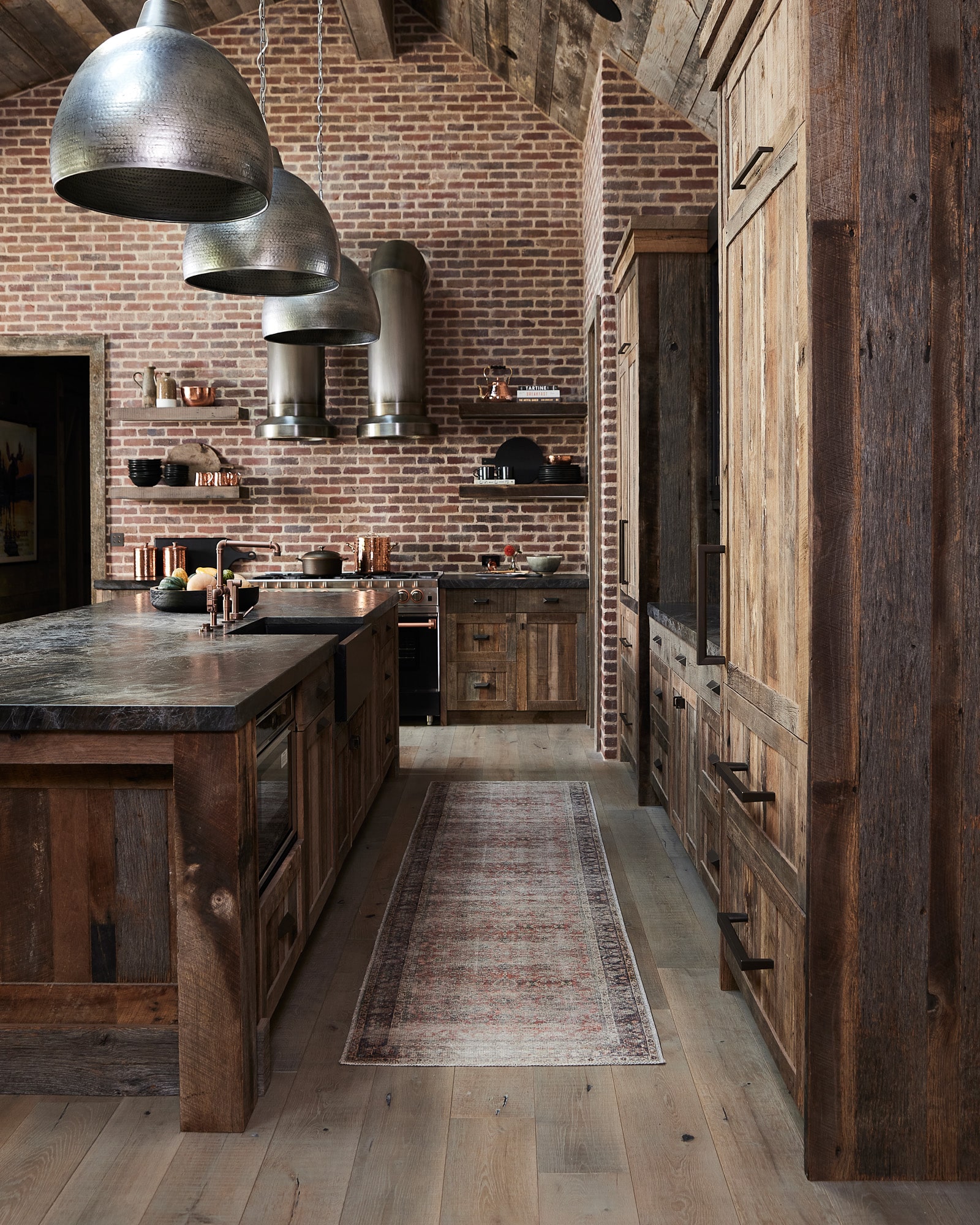 A rustic kitchen with exposed brick walls, wooden cabinetry, a large island with dark countertop, metallic pendant lights, open shelves, and a narrow patterned rug on light wooden floors—perfect for a ranch for sale or recreational land retreat.