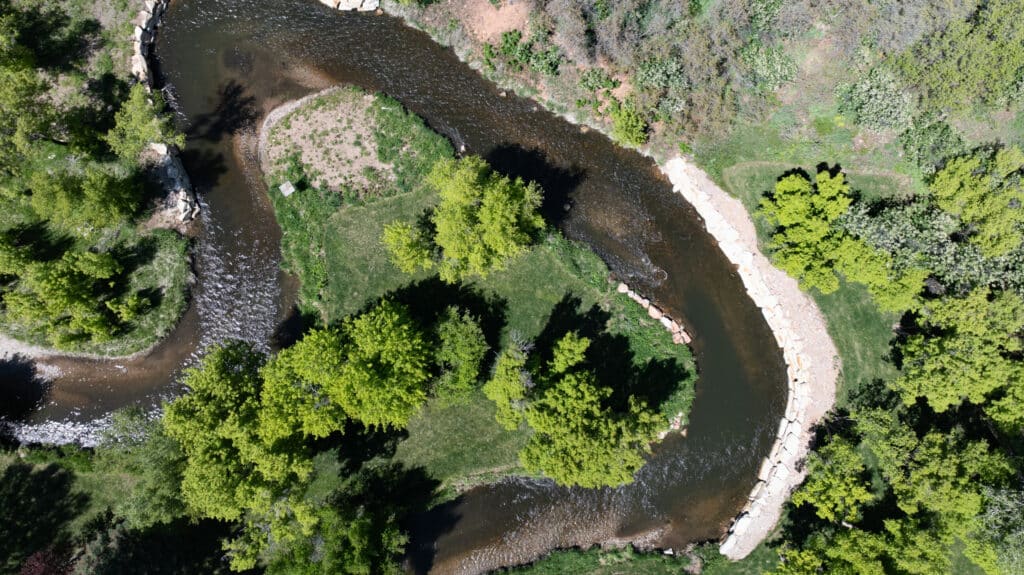 Aerial view of a winding river bordered by green trees and grassy areas, with stone retaining walls and a small dirt patch—perfect recreational land or potential cattle ranch available.