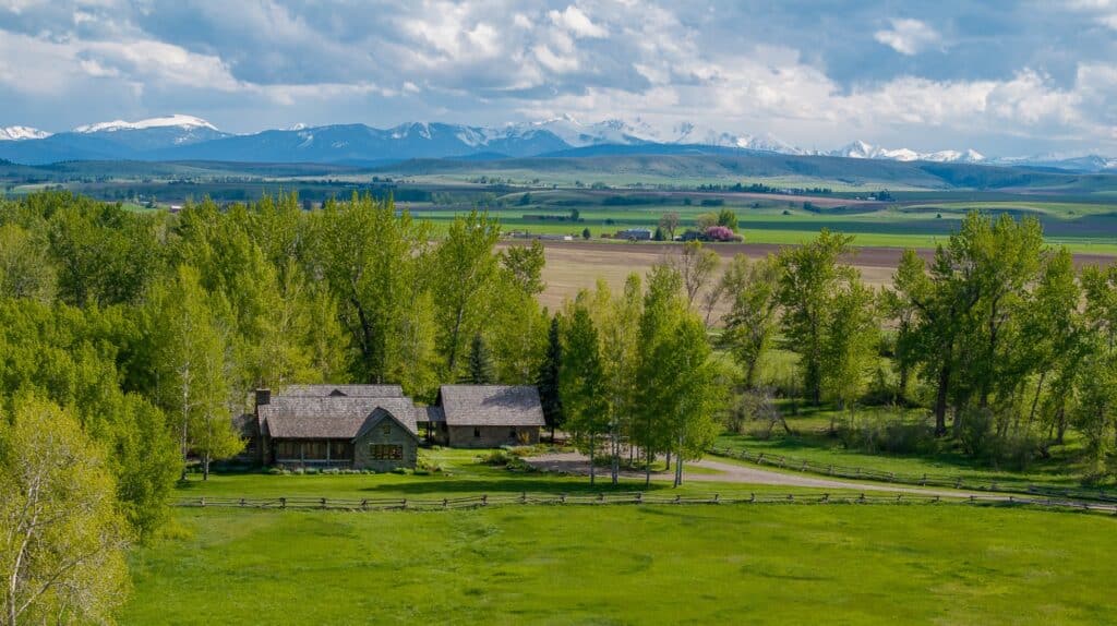 A rustic house on a cattle ranch is surrounded by green trees and fields, with a wooden fence in front and snow-capped mountains visible in the distance under a partly cloudy sky.