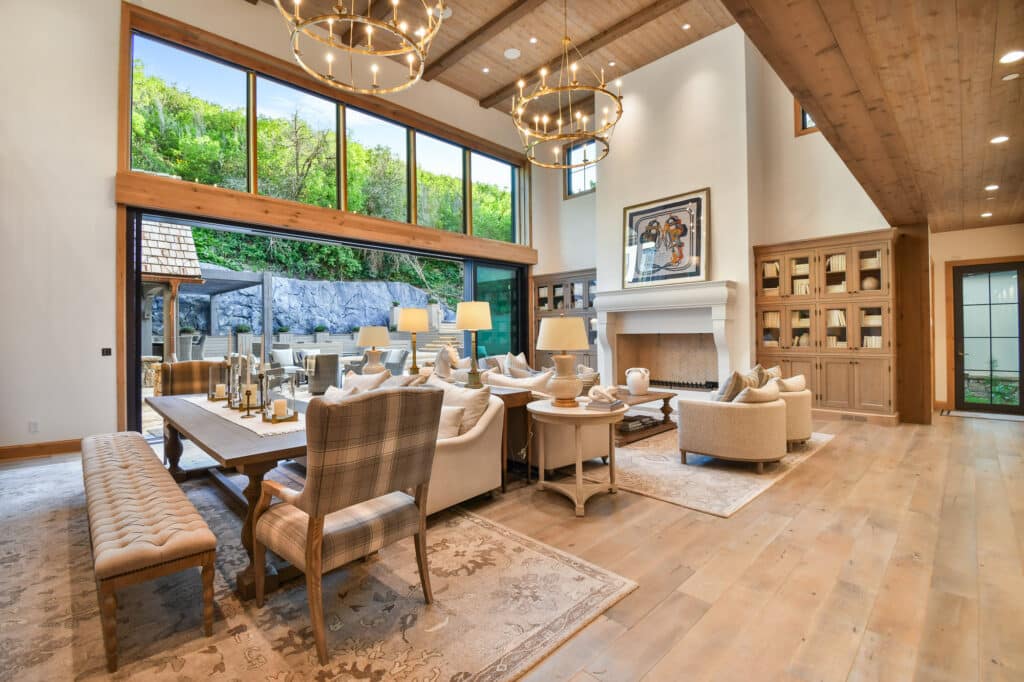 Bright, spacious living and dining area with wood floors, exposed beams, large chandeliers, neutral furniture, and glass doors opening to a patio with greenery—perfect for relaxing after a day on your cattle ranch or hunting property.