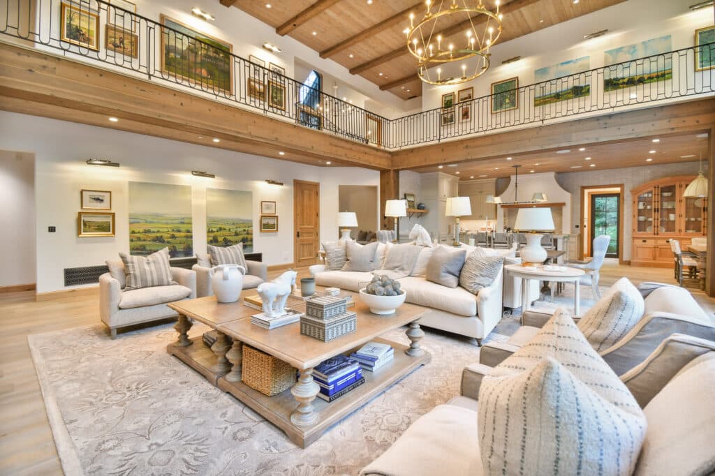 Spacious, elegant living room with beige sofas, a large rug, and a wooden coffee table. High wood ceilings and an open loft create a warm ambiance, making this ranch for sale perfect for relaxing while enjoying natural light from large windows.