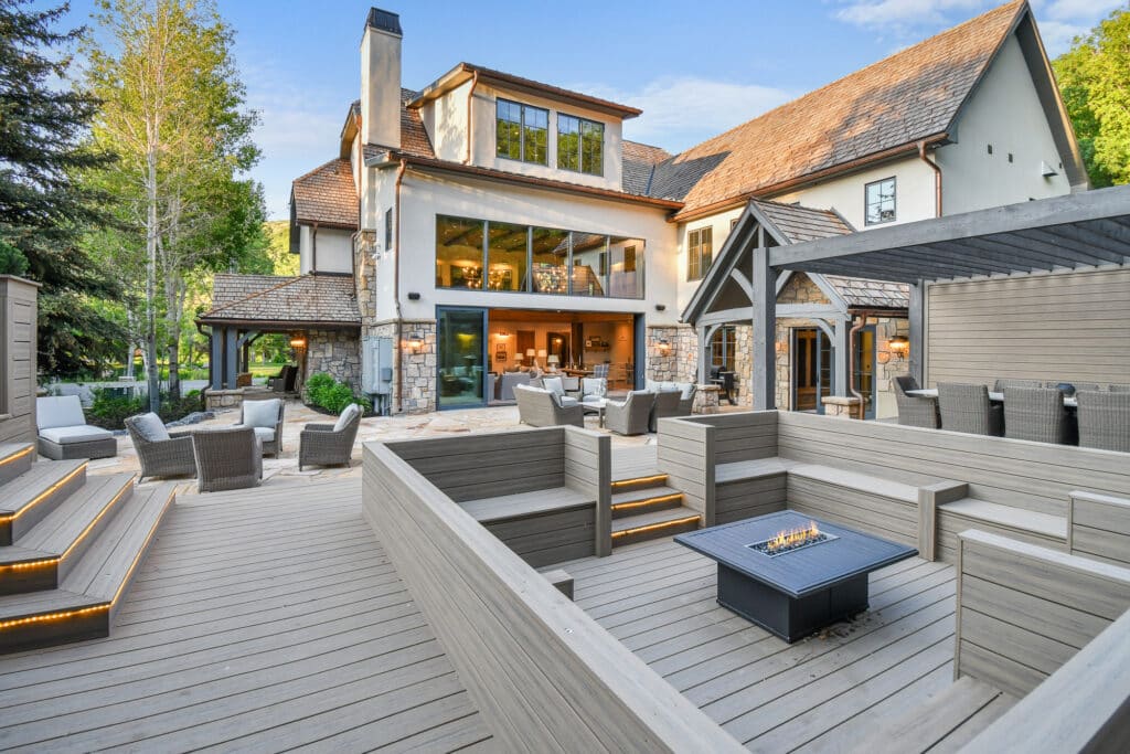 Spacious outdoor patio of a large modern house with wood decking, built-in seating, a fire pit, and patio furniture. Large windows overlook greenery and trees—perfect for enjoying life on a ranch for sale or recreational land.