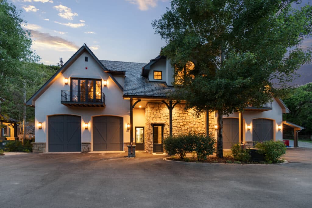 A modern house with stone and white exterior, three large garage doors, outdoor lights illuminating the entrance, and trees surrounding the driveway at dusk—ideal as a hunting property or ranch for sale.