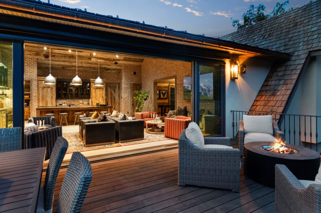 Modern patio with wicker chairs and a fire pit leads into a cozy, open living room and kitchen area with brick walls, pendant lights, and rustic wooden accents—perfect for enjoying evenings on your new ranch for sale.