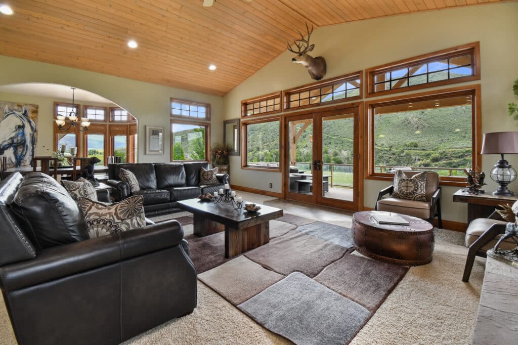 Spacious living room with black leather sofas, patterned rug, wooden coffee table, large windows showcasing green hills, mounted deer head, and vaulted wood ceiling. Perfect for a cattle ranch or hunting property; dining area and artwork visible in the background.