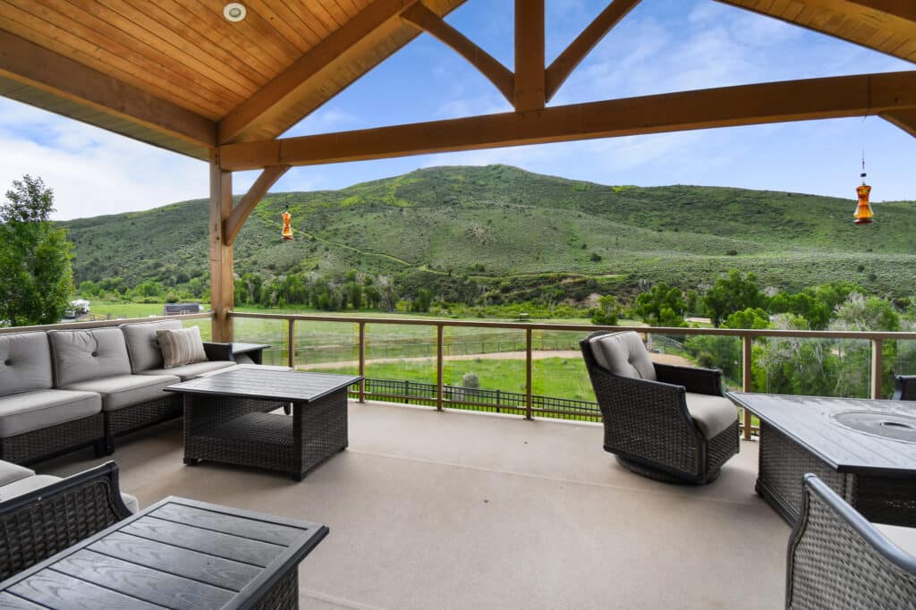 Covered patio with cushioned outdoor furniture and tables, overlooking a green, grassy hillside and scenic landscape—perfect for enjoying your ranch for sale or hunting property under a partly cloudy sky.
